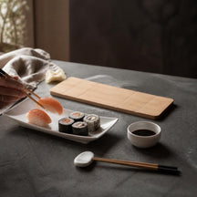 Eetstokjes pakken sushi van een wit sushibord naast dipschaaltje en plank